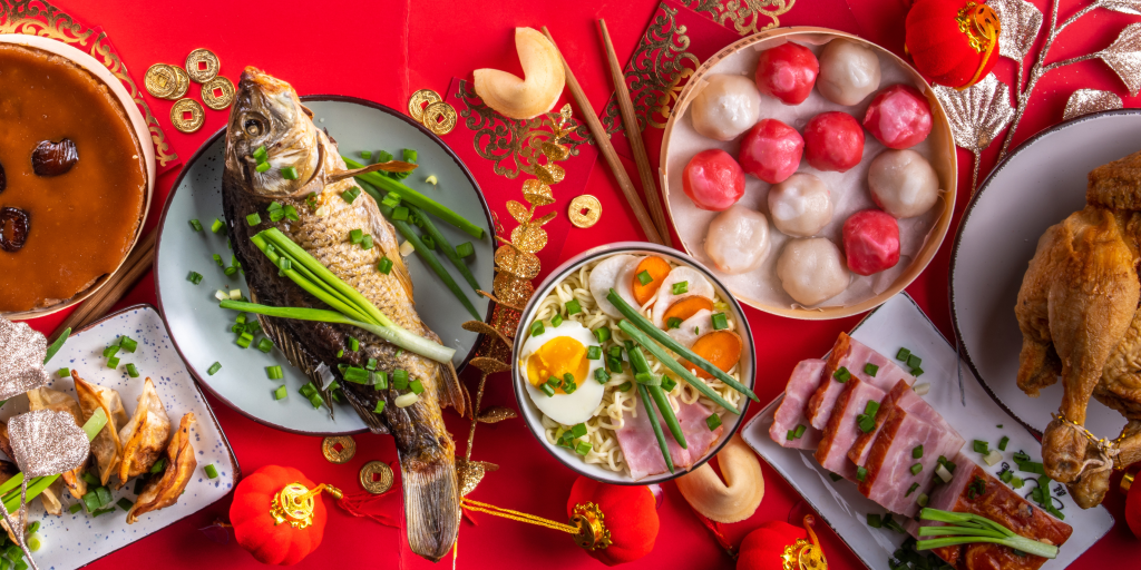 Celebrating Lunar New Year in the Classroom - the picture is of a feast featuring a whole steamed fish, noodles, steamed bao buns and dumplings on a red tablecloth with small red lanterns and gold coins.