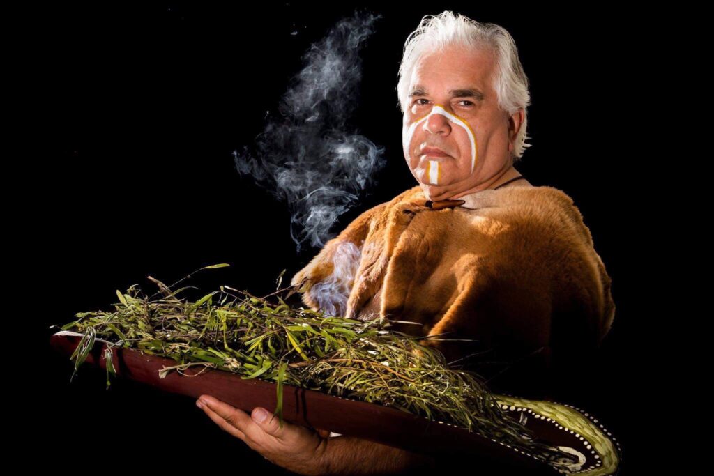 Gene Blow, proudly painted in ochre and wearing an animal-skin cloak, holds a coolamon filled with smoking gum leaves.