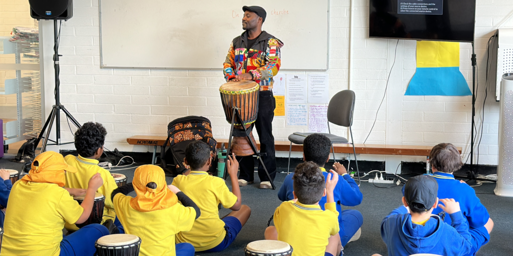 May Cultural Celebrations: Diversity, Education, and Awareness – this photo is of our Rhythms and Instruments of West Africa presenter playing the djembe with students in front of him.